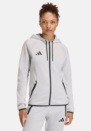 Femme portant un sweat à capuche zippé Adidas gris clair et un pantalon assorti avec fermeture éclair noire et détails du logo, debout devant un fond uni.