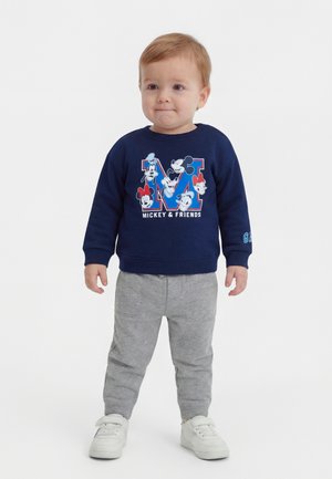 Småbarn stående iført marineblå Mickey & Friends-sweatshirt, grå bukser og hvide sneakers mod en ensfarvet hvid baggrund.