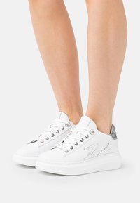 Hvite sneakers med en glatt lærøvre, rund tå, sølv logo detaljering og teksturerte bakakenter. Litt hevet gummisåle.