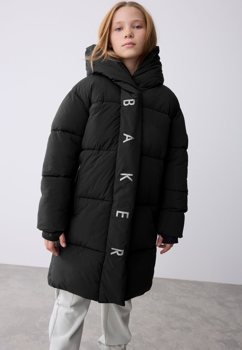 Zwarte donzen jas met capuchon, voorzien van doorgestikte secties en het woord "BAKER" in grijs op de voorkant. Draagt lichtgekleurde broek.