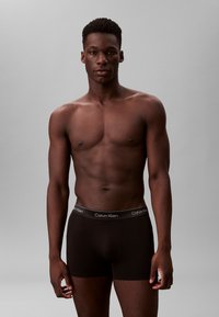 Schwarze Baumwoll-Boxershorts mit einem grauen Bund, auf dem das „Calvin Klein“-Logo zu sehen ist, präsentiert an einem männlichen Modell vor einem neutralen Hintergrund.