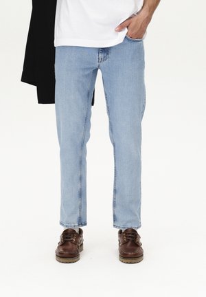 Jeans de pierna recta en azul claro con un acabado desgastado, combinados con zapatos marrones de cordones. Se lleva una camiseta blanca debajo de una chaqueta negra.