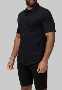 Chemise noire à manches courtes et à boutons en matériau texturé, dotée d'une poche poitrine, de boutons contrastants et associée à un short noir.