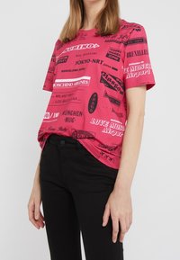 Růžové tričko s krátkým rukávem zdobené černými grafikami a texty souvisejícími s cestováním, volného střihu a z měkké bavlny. V kombinaci s černými kalhotami.