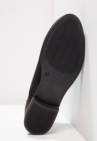 Suela de zapato de ante negro con una base de goma texturizada que presenta patrones de ondas y una etiqueta de tamaño impresa. Bordes suaves con una forma redondeada.