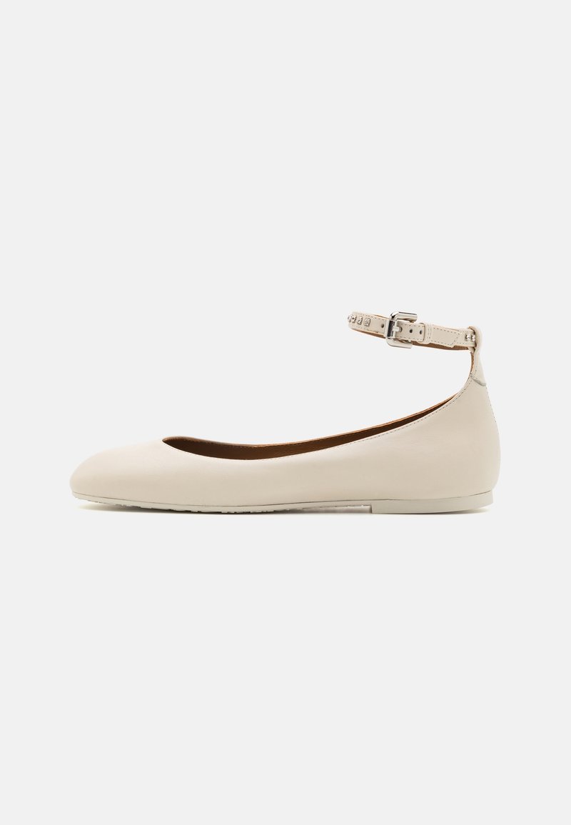 Ballerines plates beiges avec une sangle de cheville ornée de petits clous en argent et d'une boucle argentée, présentées sur un fond blanc uni.