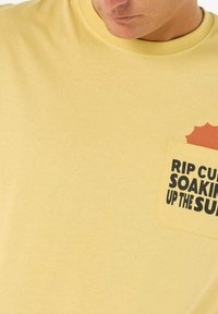 Gul bomulds T-shirt med en lomme og et grafisk tryk, der står "RIP CURL SOAKING UP THE SUN" i sort skrift. Rund halsudskæring.