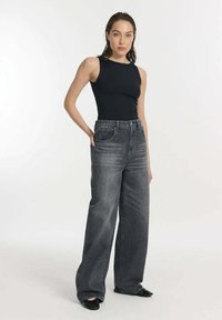 Zwarte mouwloze top gecombineerd met hoogwaist, wijdvallende donkergrijze jeans. Het model staat in een neutrale pose en toont de pasvorm van de outfit.