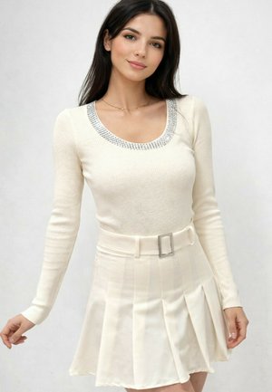 Femme aux longs cheveux noirs portant un pull blanc côtelé avec un col orné de strass et une jupe plissée blanche avec ceinture, posant devant un fond uni.