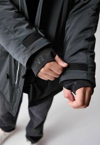 Melna ūdensizturīga jaka ar gludu tekstūru, ar Velcro aprocēm un rāvējslēdzēja aizdari, uzvilkta virs tumša garām piedurknēm krekla.