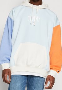 Felpa azzurra chiara con maniche arancioni e blu, presenta un logo bianco "TOMMY" sul petto e una tasca frontale; realizzata in tessuto morbido.