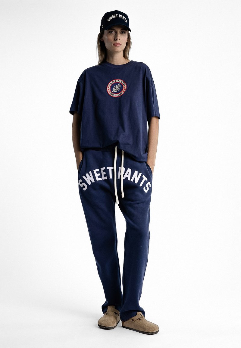 T-shirt oversize bleu marine avec un graphique circulaire, pantalon de survêtement bleu marine assorti avec l'inscription "SWEET PANTS" en blanc, et une casquette noire avec une inscription blanche.