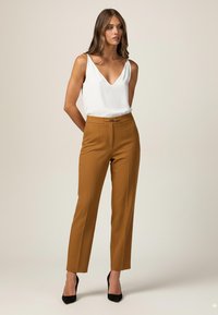 Brune skræddersyede bukser med en tekstureret finish, høj talje og en metalaccent ved lukningen, parret med en hvid V-hals tanktop.