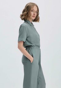 Kurzarmpullover aus weicher, sanfter Tealfarbe, mit knöpfbarem Frontbereich, elastischem Taillenbund und seitlichen Taschen für Funktionalität.