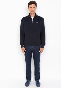 Marineblauer Pullover mit Reißverschluss-Kragen aus weichem Stoff. Getragen mit dunkler Jeans und blauen Sneakers. Einfaches Design, keine sichtbaren Muster oder Akzente.