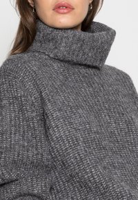 Kvinna som bär en tjock grå stickad polotröja med ribbat mönster, visar sina läppar och en del av sitt hår mot en heltäckande bakgrund.