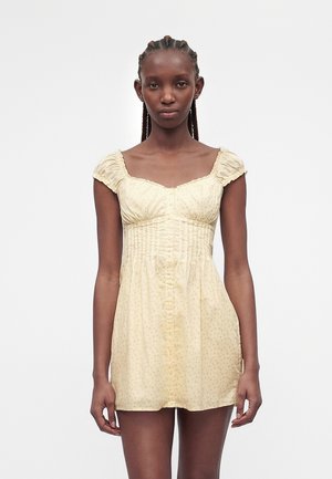 Schlanke Frau mit geflochtenem Haar, die ein kurzes, hellgelbes, knopfbares Sommerkleid mit kurzen Puffärmeln vor einem einfarbigen weißen Hintergrund trägt.