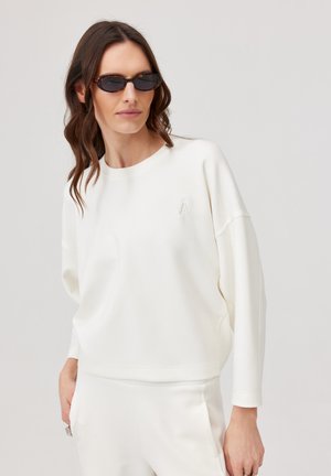 Kobieta o falowanych brązowych włosach, nosząca ciemne owalne okulary przeciwsłoneczne oraz luźną białą bluzkę z długimi rękawami i dopasowane białe spodnie, pozująca na jasnym tle.