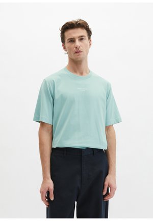 Man draagt een lichtblauw T-shirt met korte mouwen, voorzien van een klein wit logo, en zwarte broek, staand tegen een effen witte achtergrond.