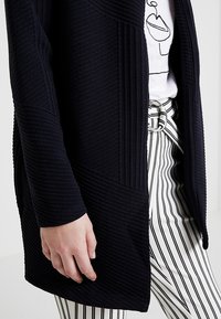 Svart texturerad kofta med ribbat mönster och långa ärmar, bärs över en vit skjorta och randiga byxor med bältesdetalj.