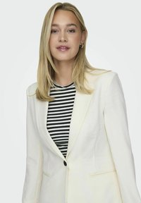 Hvid blazer med klapkraver, enkelt sort knap og to frontlommer. Båret over en sort og hvid stribet bluse. Glat tekstur.