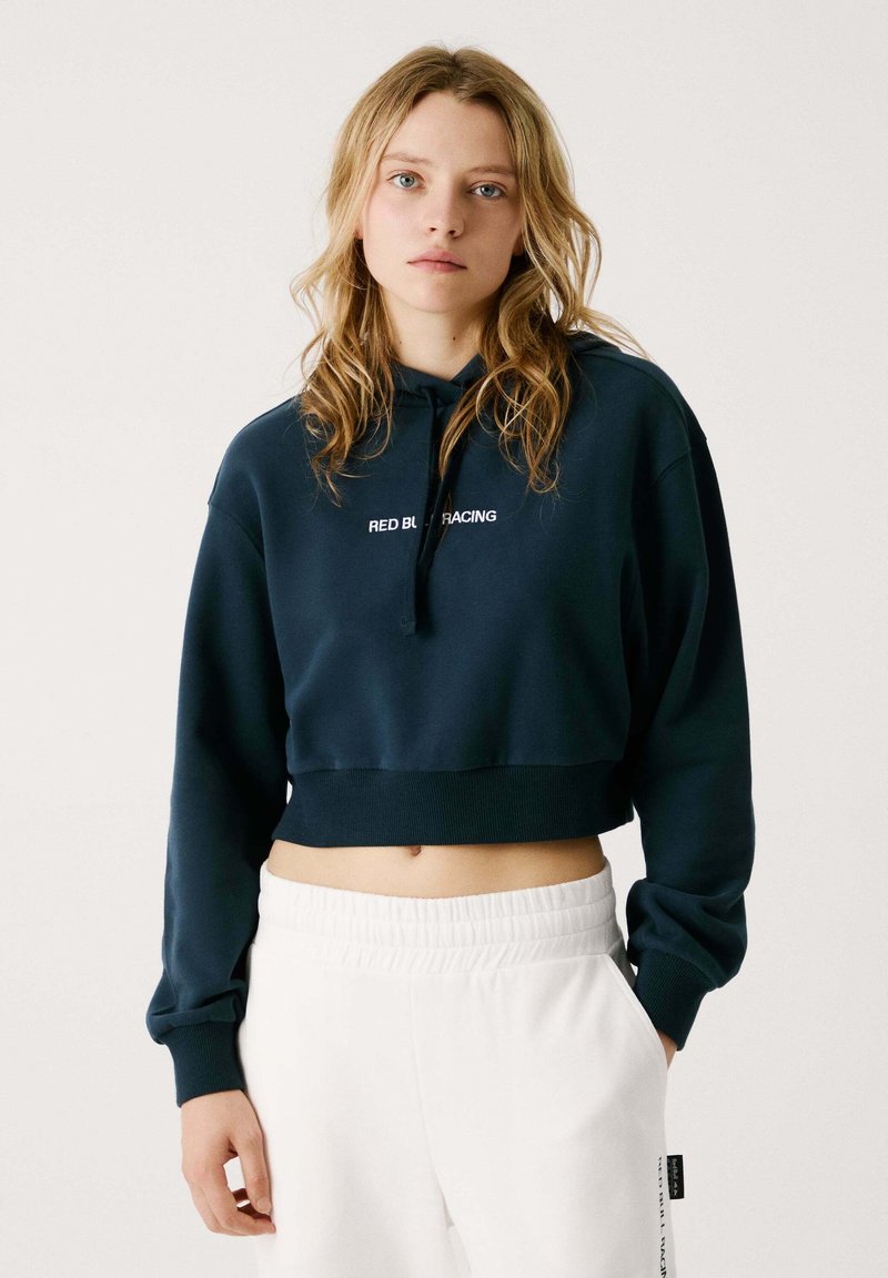 Mladá žena s voľnými blond vlasmi, oblečená v námorníckej modrej skrátenej mikine s nápisom "RED BULL RACING" a bielych elastických nohaviciach s vysokým pásom.