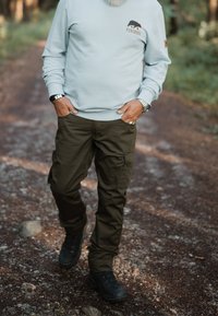 Ljusblå sweatshirt med logotyp på bröstet, olivgröna cargobyxor och svarta vandringskängor, går på en grusväg i ett skogsområde.