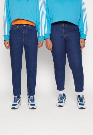Två individer bär blåa sweatshirts och mörkblå jeans. Båda har vita och blå sneakers. Jeansen har en figurnära design med uppvikta fållar.