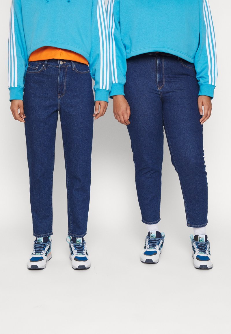 Dos personas llevan sudaderas azules y jeans azul oscuro. Ambas tienen zapatillas blancas y azules. Los jeans tienen un diseño ajustado con dobladillos enrollados.