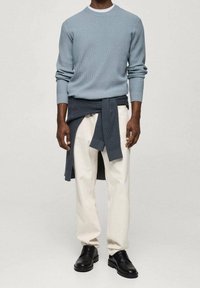 Homme portant un pull côtelé bleu clair, un pantalon blanc, des chaussures de ville noires, avec un pull foncé noué autour de la taille, debout devant un fond uni.