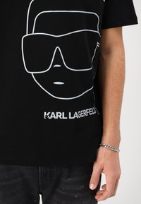 Black t-shirt featuring a large white outline of sunglasses and a face design. "KARL LAGERFELD" in bold white text at the bottom.