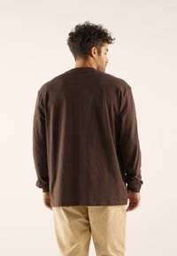 Chemise brune à manches longues en coton, avec un col rond et une coupe décontractée. Associée à un pantalon de couleur claire. Texture simple et lisse.