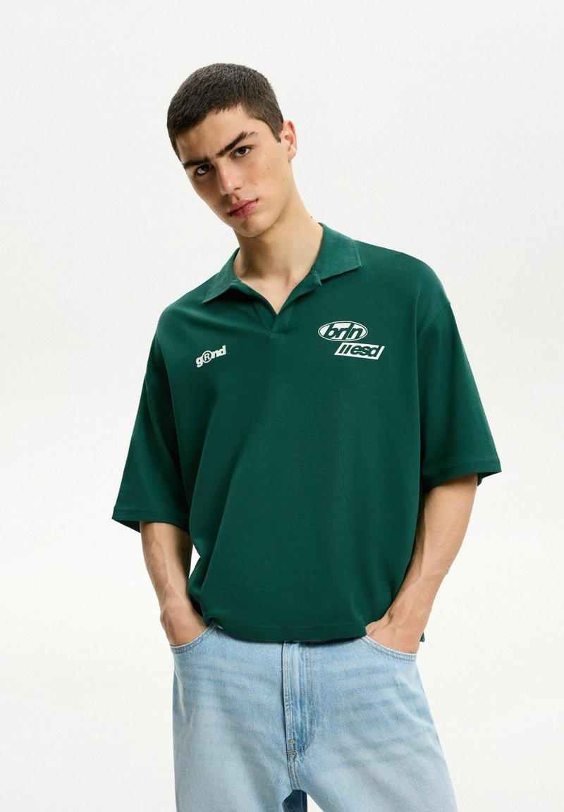 Mörkgrön pikétröja med kort krage, prydd med vita logotyper på vänster bröst och ärm, ihop med ljusblå jeans.