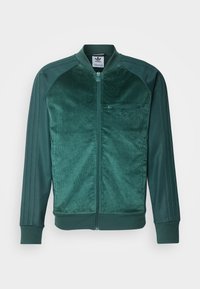 Grön kordjacka med dragkedja framtill, ribbade ärmslut och tre ränder längs ärmarna. Har en dragkedjeficka på vänster bröst.