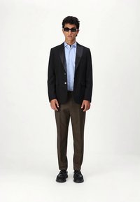 Must täisklassikaline jakk satiinist revääridega, heleroheline särk, pruunid püksid ja mustad kingad. Modell kannab tumedaid päikeseprille, seisab valgel taustal.