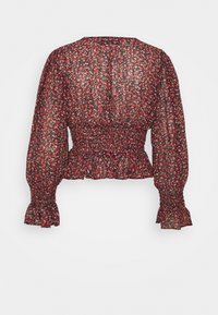 Blouse à fleurs rouges avec des manches longues, taille froncée, tissu transparent texturé, poignets volantés et petits motifs floraux sur fond noir.