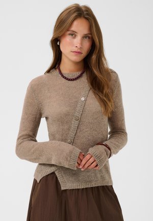Jeune femme aux longs cheveux bruns porte un pull beige asymétrique boutonné, une jupe marron, un collier de perles violet, des boucles d'oreilles en perles et un bracelet marron.