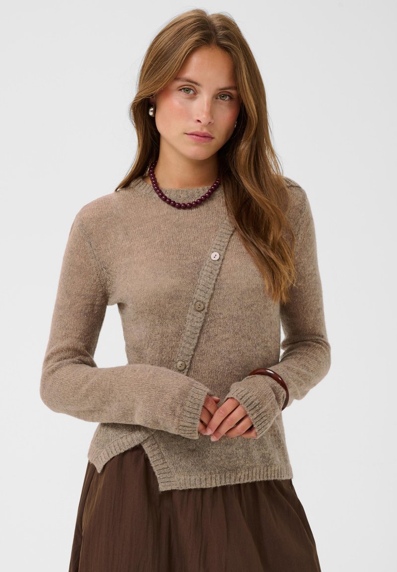 Jeune femme aux longs cheveux bruns porte un pull beige asymétrique boutonné, une jupe marron, un collier de perles violet, des boucles d'oreilles en perles et un bracelet marron.