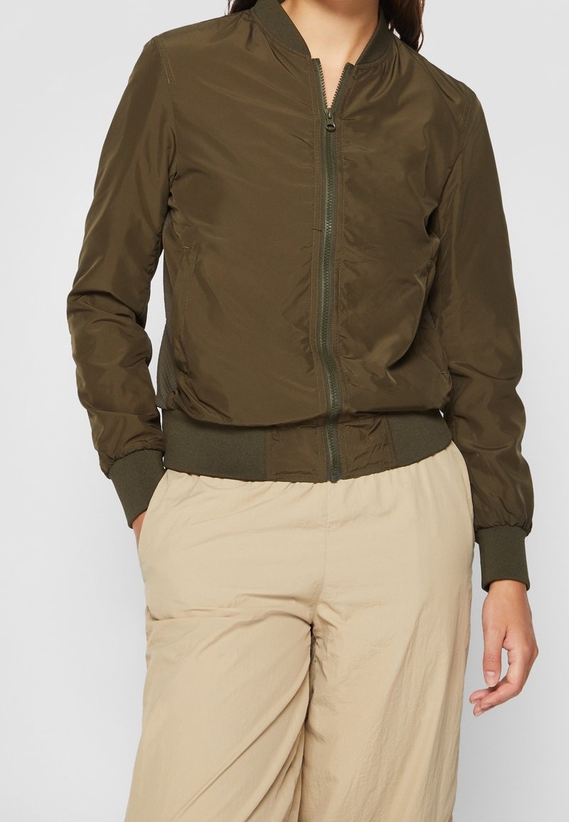 Olivgrön bomberjacka i slät tyg, med dragkedja framtill, ribbade ärmslut och elastisk nederkant, tillsammans med ljusbeige lösa byxor.