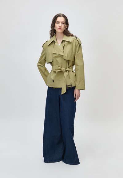 Γυναίκα με σγουρά μαλλιά, φορώντας μπουφάν ελιάς με ζώνη και φαρδιά σκούρα μπλε τζιν, στέκεται μπροστά σε απλό φωτεινό φόντο.