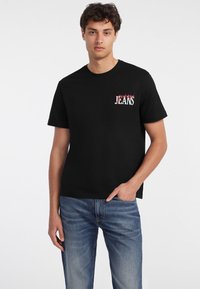 Melns kokvilnas T-krekls ar sarkano un balto logo drukātu kreisajā krūtīs, apvienots ar gaiši ziliem džinsu biksēm.