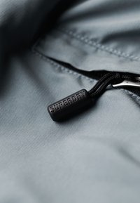 Grijze stof met een ritssluiting en een zwarte rubberen trekknop met de opdruk "SUPERDRY." Gladde textuur en eenvoudig ontwerp.