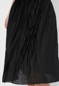 Gonna nera plissettata con un design testurizzato e fluido. Presenta una vita arriccita e si estende fino a metà polpaccio. Tessuto leggero e sottile.