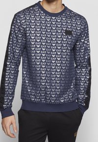 Sudadera azul marino con un patrón de logo EA7 en toda la prenda, cuello y puños de punto, y detalles negros en las mangas. Textura suave.