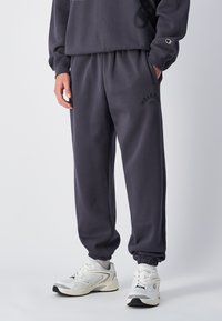 Un jogging gris foncé avec une coupe décontractée, une taille élastique, des poches latérales et un imprimé logo noir. Associé à des baskets blanches.