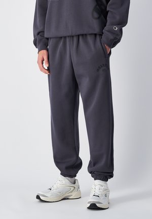 Dunkelgraue Jogginghosen mit lockerer Passform, elastischem Bund, seitlichen Taschen und schwarzem Logo-Druck. Kombiniert mit weißen Sportschuhen.