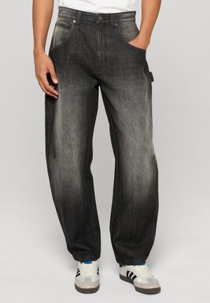 Laza fekete farmernadrág, kifakult mosással, nagy elülső zsebekkel és felhajtott szegéllyel. Fehér és szürke sportcipővel viselve.