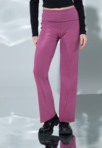 Roze ribgebreide wijde broek met een dikke, gestructureerde tailleband. Gecombineerd met zwarte chunky plateau-laarzen, tegen een reflecterende achtergrond.