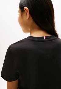 T-shirt en coton noir avec un col rond, des manches courtes, un tissu texturé et un petit accent décoratif rouge, blanc et bleu au dos du col.