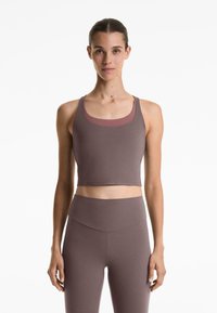 Kvinna som står framåtvänd, iklädd en mauve crop tank top och högmidjade leggings mot en vit bakgrund.